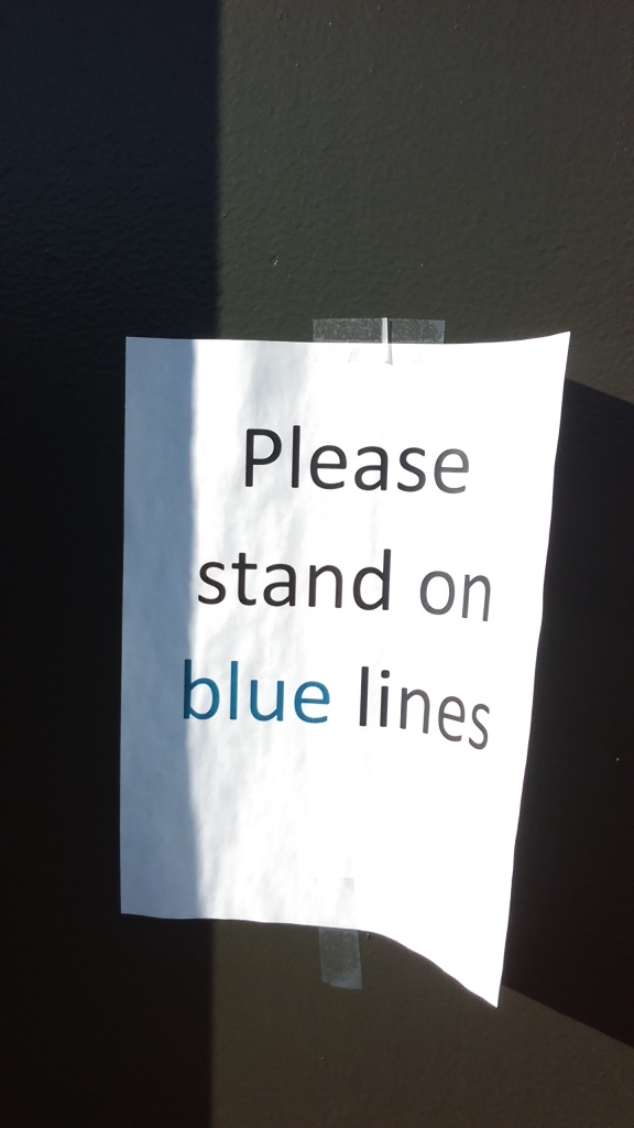 A sign printed on white paper and taped to a pole reads, "Please stand on blue lines," and the words are printed in black except the word blue, which is printed in blue.