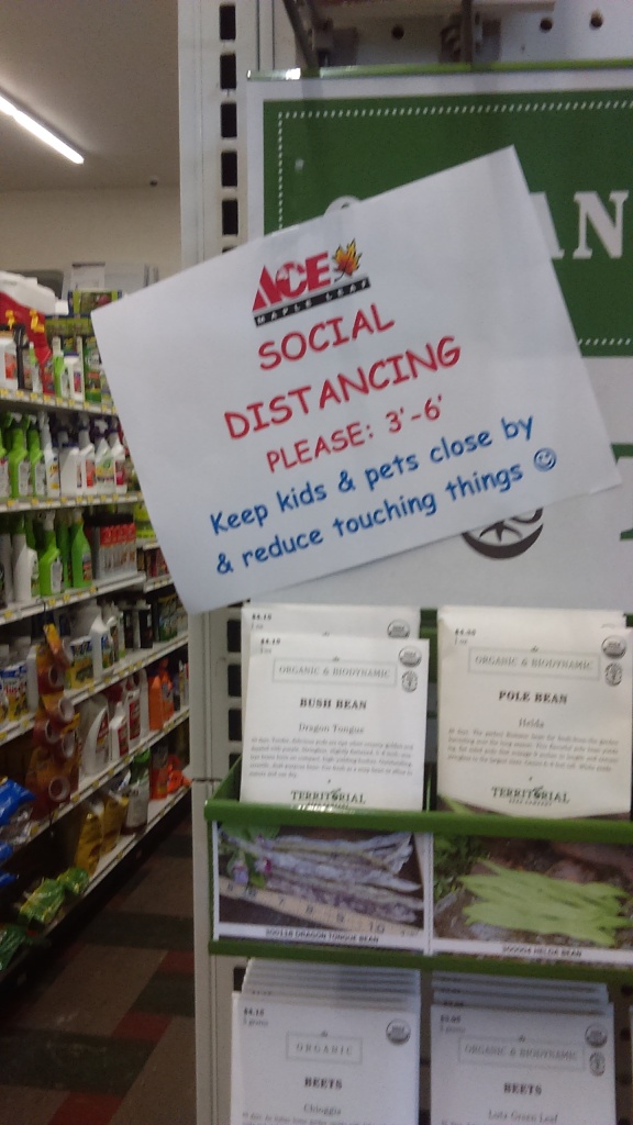 On an end cap of an aisle with food seeds, a sign sits haphazardly above the seeds. The top of the sign shows the ACE Maple Leaf hardware store logo. Under that in red text and all caps, it reads, "SOCIAL DISTANCING PLEASE: 3'-6'" and below that is blue text that reads, "Keep kids & pets close by & reduce touching things" followed by a blue smiley face.