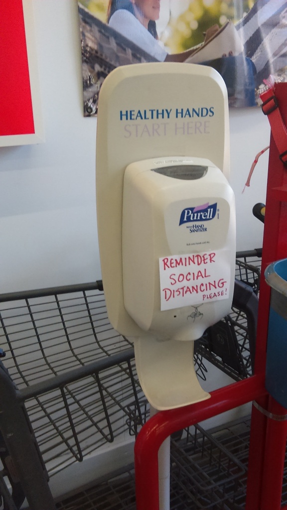 A Purell-brand hand sanitizer station has a hand-written sign taped to it. Red letters on white paper in all caps read, "REMINDER SOCIAL DISTANCING" with "PLEASE!" written smaller at the bottom, perhaps as an afterthought.