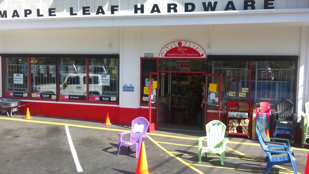 ACE Maple Leaf Hardware storefront. Both doors to the entrance are open. An employee stands just inside. A few feet outside of the door are two big plastic lawn chairs with signs on white paper taped to them with blue painter's tape. Between the chairs and a little further away from the doors is a traffic cone.