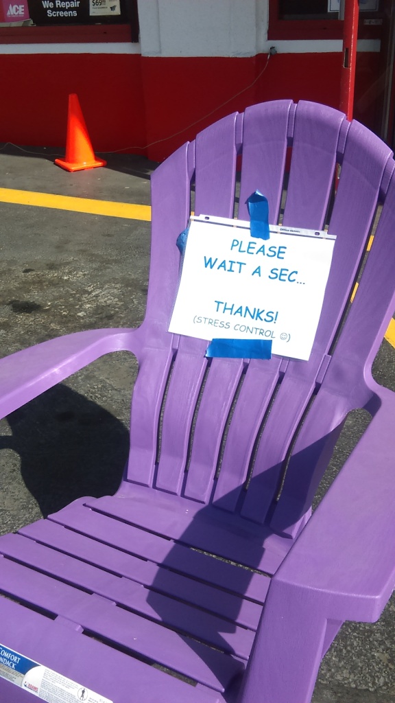 A sign is taped to a lawn chair outside a hardware store. The first part reads in all caps, "PLEASE WAIT A SEC" followed by three periods. There is a line left blank, and then it says "THANKS!" in all caps. Under that is a parenthetical phrase that reads "STRESS CONTROL" in all caps and is followed by a smiley face inside the parentheses.