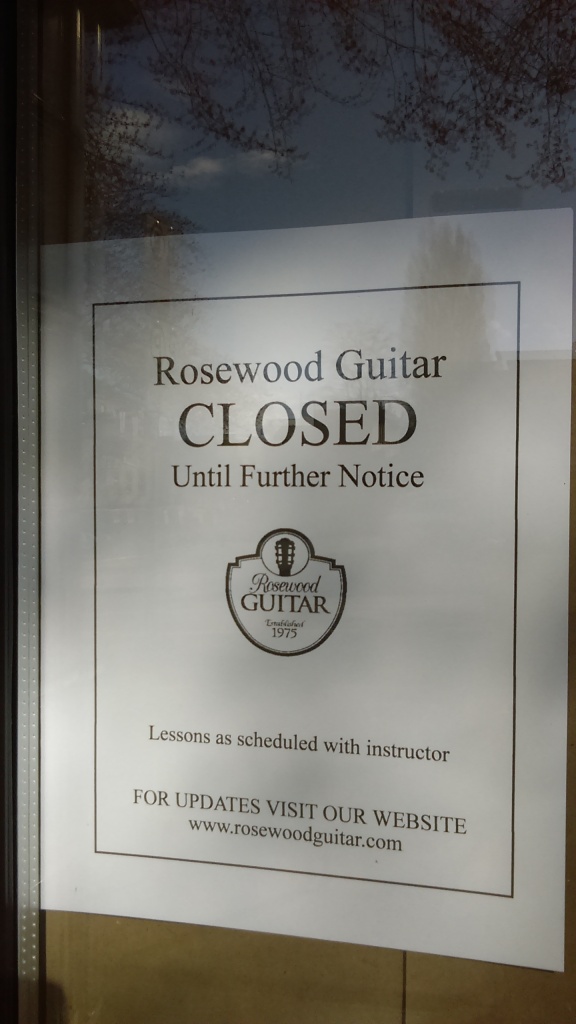 March 23, 2020, Greenwood, Seattle. A sign on the door of Rosewood Guitar bears the name and crest of the store, and reads, "CLOSED until further notice." The bottom of the sign reads, "Lessons as scheduled with instructor," and, "FOR UPDATES VISIT OUR WEBSITE" and has the web site www.rosewoodguitar.com listed below.