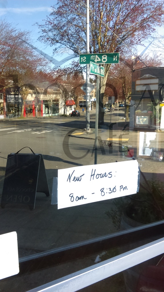 Aside from the street reflected in the window, this image shows a hand-written sign that reads "New Hours: 8am - 8:30pm" taped to the window under Chocolati Cafe's name.