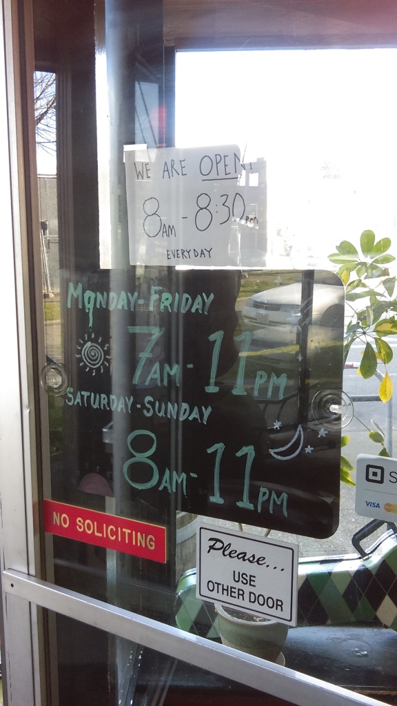 March 23, 2020, Greenwood, Seattle. The door to Chocolati is plastered with signs such as "NO SOLICITING" and "Please...USE OTHER DOOR." A big black sign with neatly handwritten green chalk reads, "MONDAY-FRIDAY 7AM-11PM," and "SATURDAY-SUNDAY 8AM-11PM" and is decorated with a sun and moon drawn in yellow. These are clearly the store's standard hours. A handwritten sign on white paper taped above it says, "WE ARE OPEN 8:00AM - 8:30PM EVERYDAY."