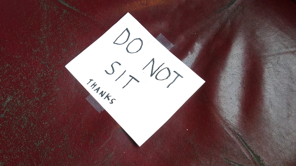 March 23, 2020, Greenwood, Seattle. Two red leather chairs inside the barista bar area of Chocolati Cafe. Each one has the same hand-written sign on white paper taped to the seat as shown here. The sign reads, "DO NOT SIT" in big letters, with "THANKS" in tiny letters at the bottom.