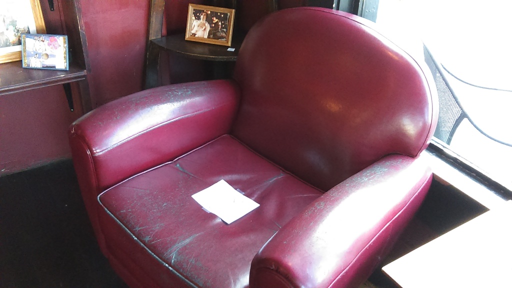 March 23, 2020, Greenwood, Seattle. One of two red leather chairs inside the barista bar area of Chocolati Cafe. It has a sign taped to the seat. The image below this one is a close up that shows what it says.